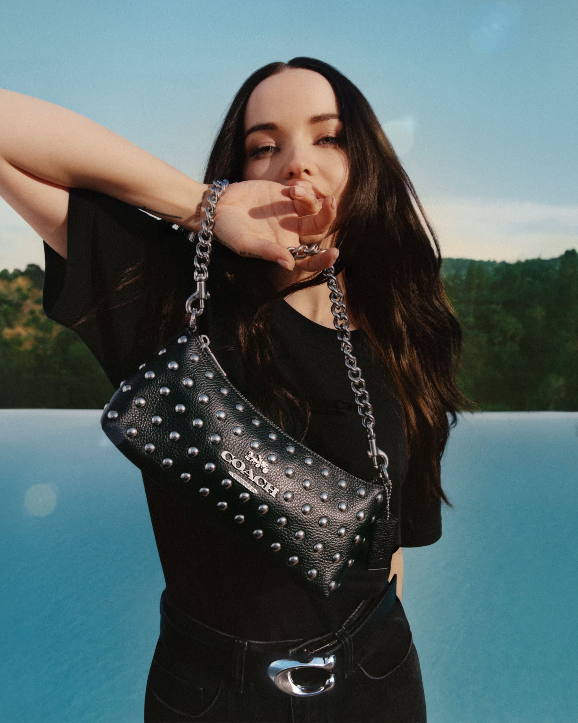 A brunette poses with a black, studded leather bag with silver chain strap against an outdoor backdrop and light blue sky.