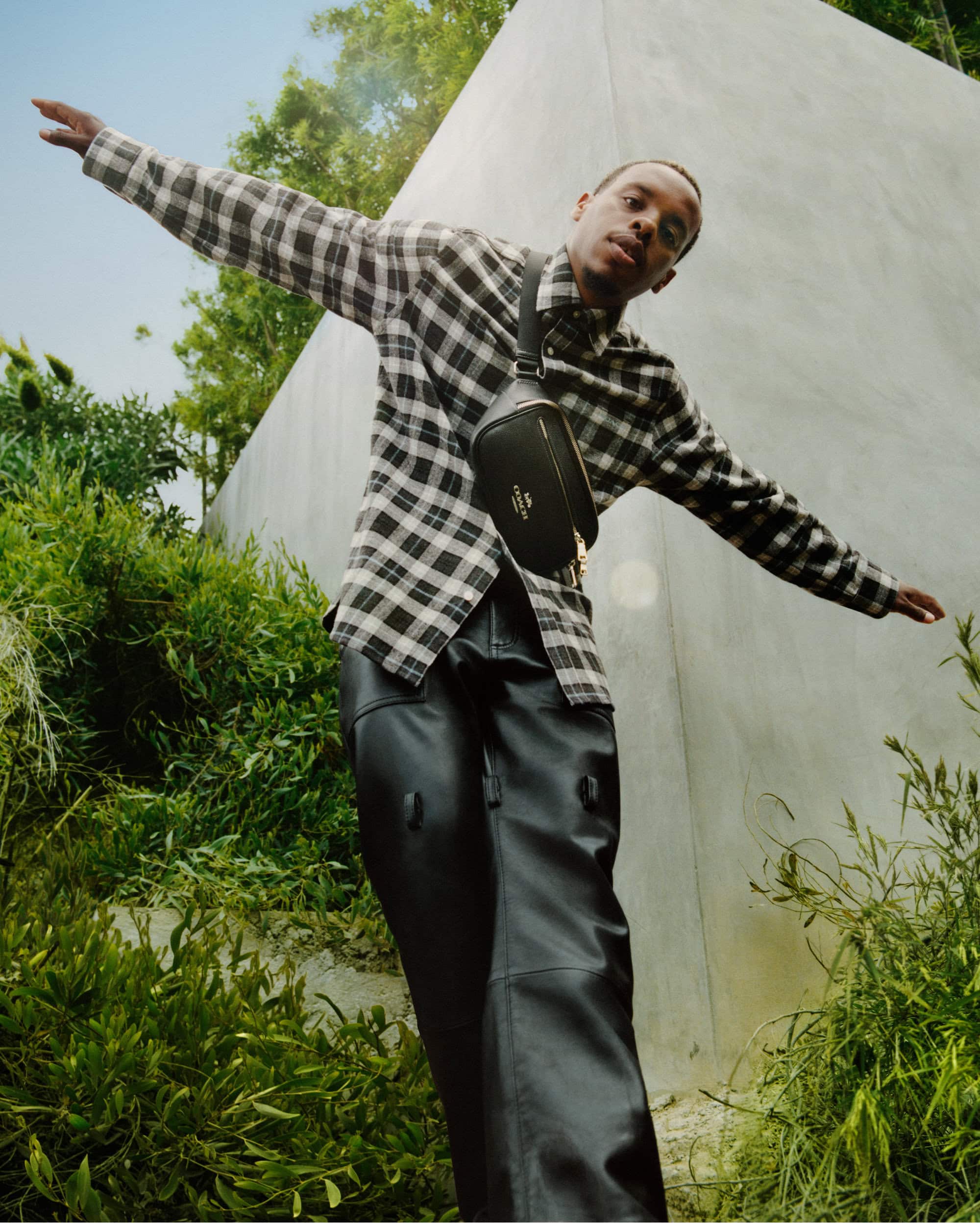 Adult male in a black and white checkered shirt, black pants, and a black crossbody bag poses outdoors with greenery and a concrete wall.