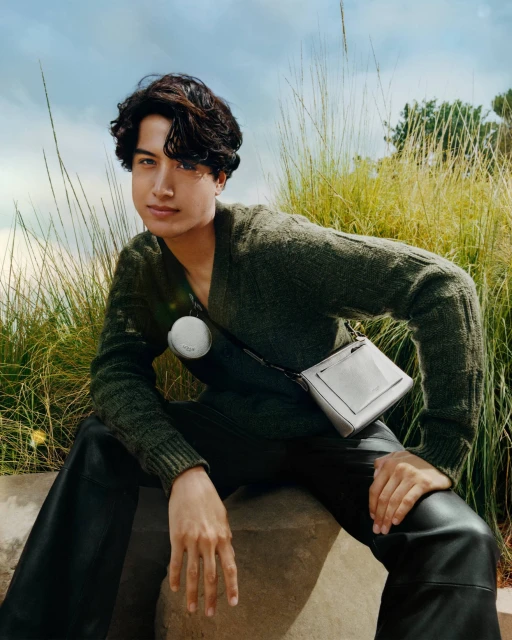 Portrait of an adult male model in an olive sweater and black leather pants with a white purse outdoors in front of foliage.