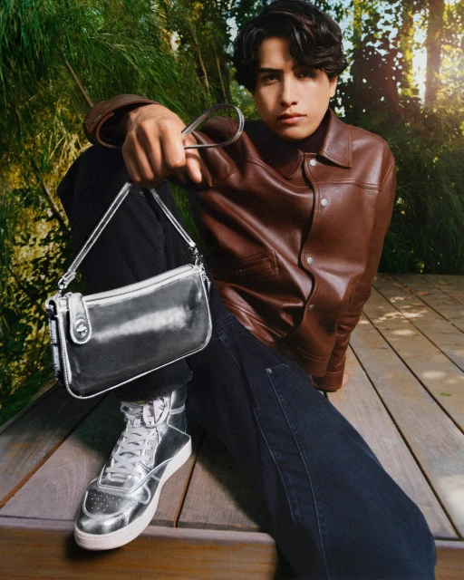 Stylish adult male poses outdoors on a wooden platform showcasing fashion sneakers and a metallic silver shoulder bag with trees behind him.