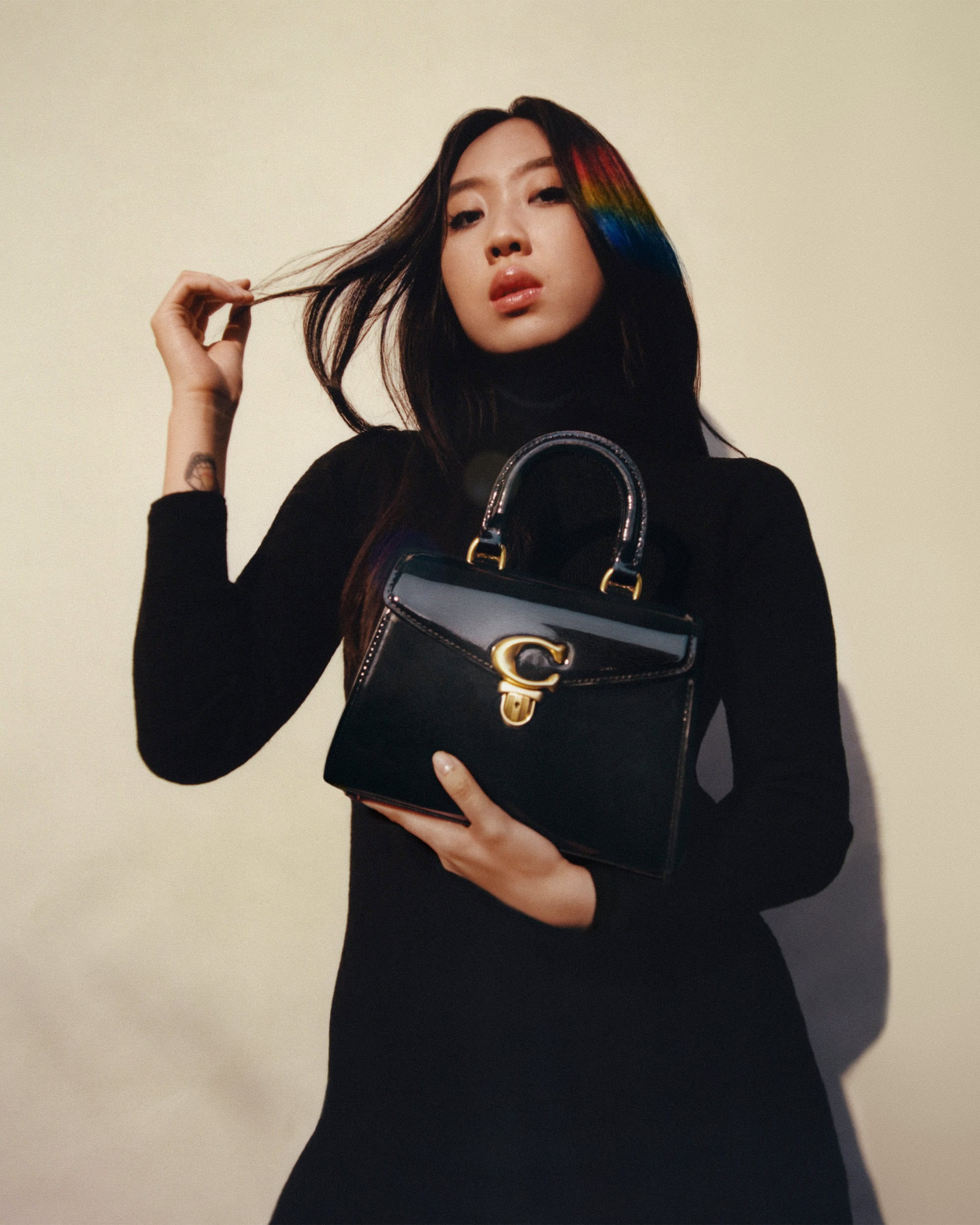 Adult female poses indoors holding a glossy black Coach handbag. She is wearing a long-sleeved black turtleneck dress with rainbow-dyed hair.