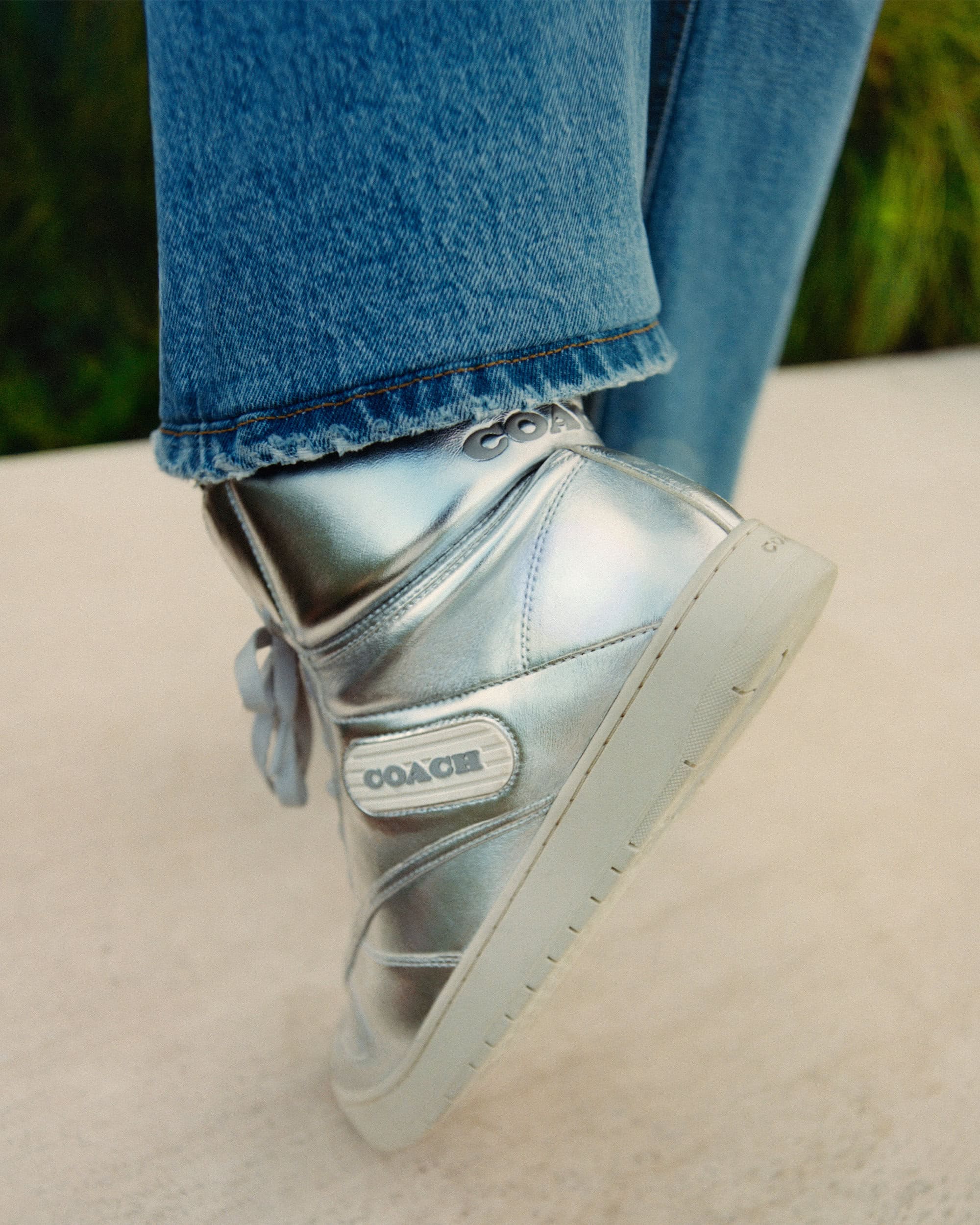 Close-up studio shot of a silver Coach high-top sneaker with blue denim jeans, displayed against a neutral background with plant life.