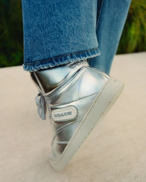 Close-up studio shot of a silver Coach high-top sneaker with blue denim jeans, displayed against a neutral background with plant life.
