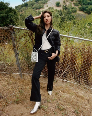 Woman poses in black puffer jacket, jeans, and metallic boots outdoors near a fence, creating a fashion-forward editorial image.