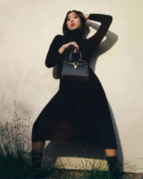 Woman in a black dress holding a black handbag against a light wall with tall grass in the foreground.
