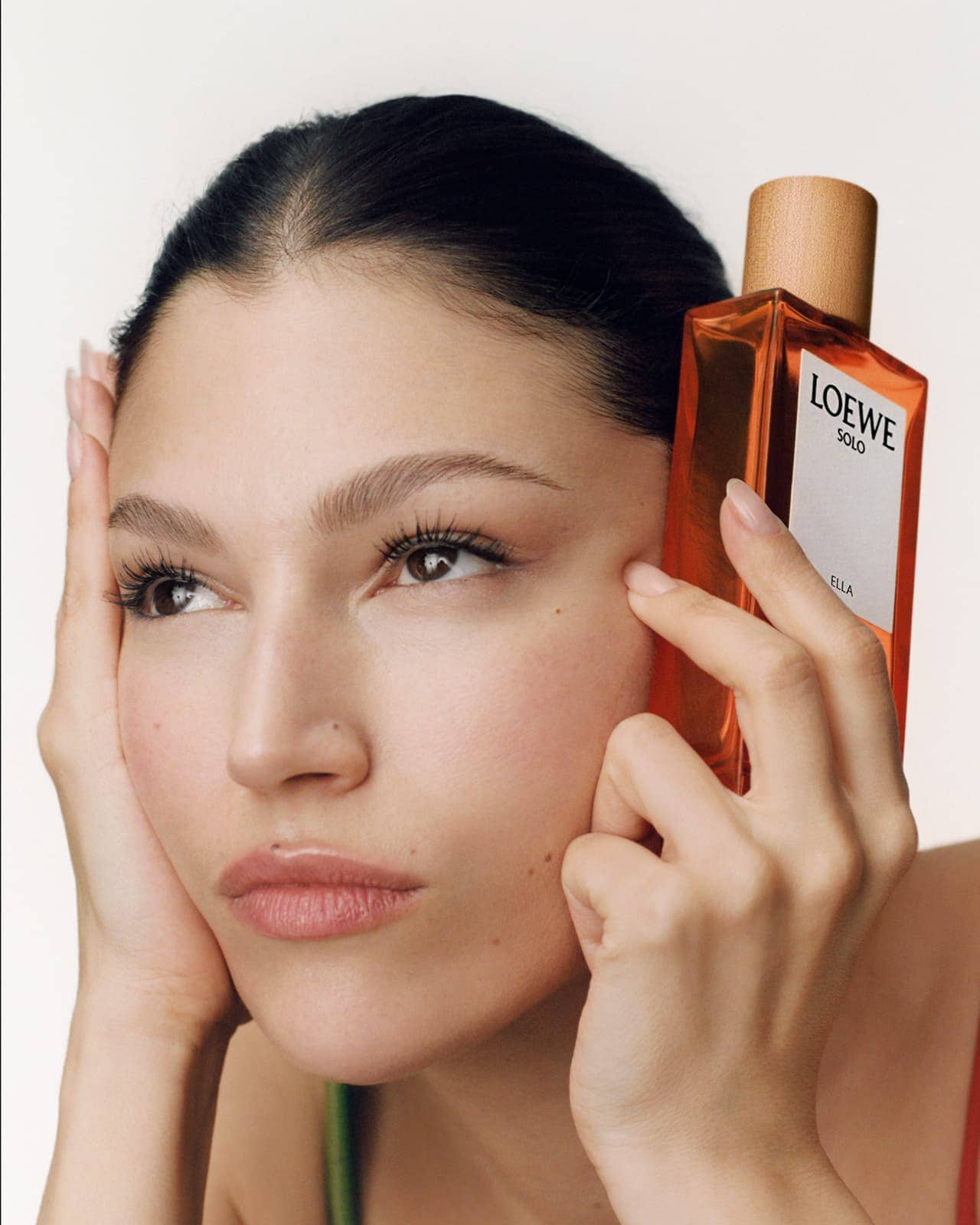 Close-up of a woman holding Loewe Solo Ella perfume bottle against her face, showcasing beauty and fragrance in a minimalist setting.