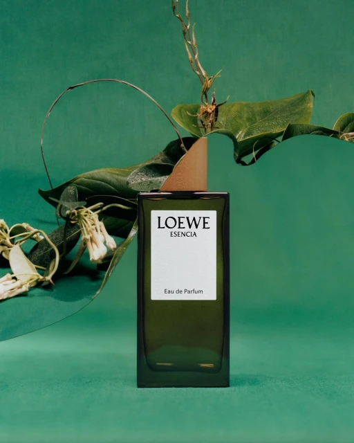 Loewe Esencia Eau de Parfum stands tall amidst green leaves and stems against a mottled green background, highlighting fragrance and nature.