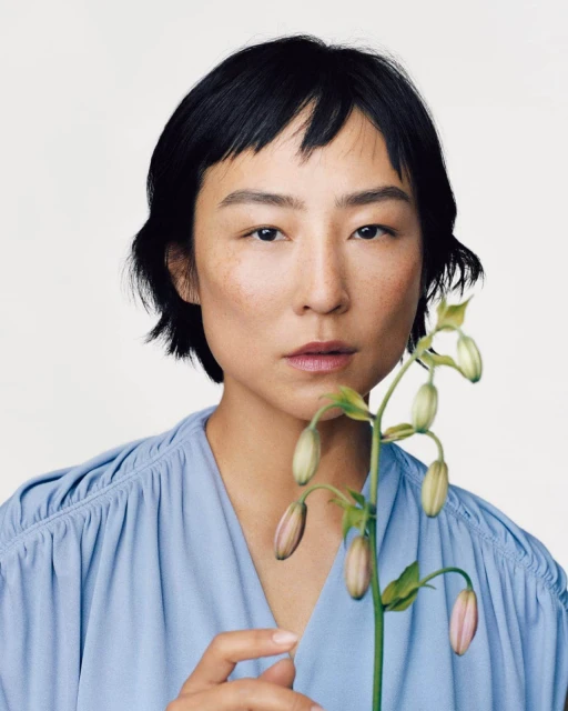 Portrait of an adult woman with short dark hair in a blue top, holding a budding flower against a white background in a photo studio.
