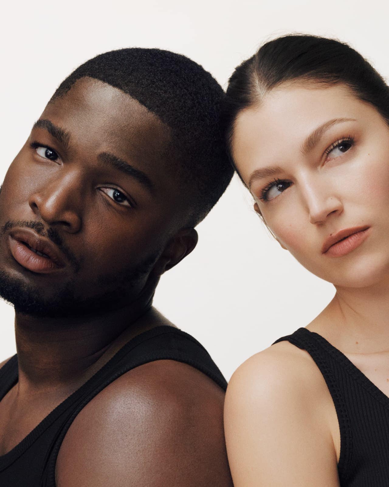 Portrait of a Black man and a White woman with their heads close together in a studio setting, both wearing black tank tops.
