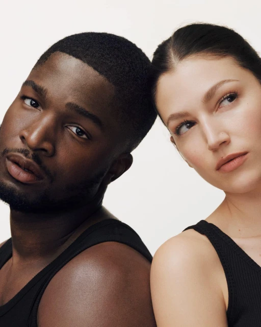 Portrait of a Black man and a White woman with their heads close together in a studio setting, both wearing black tank tops.