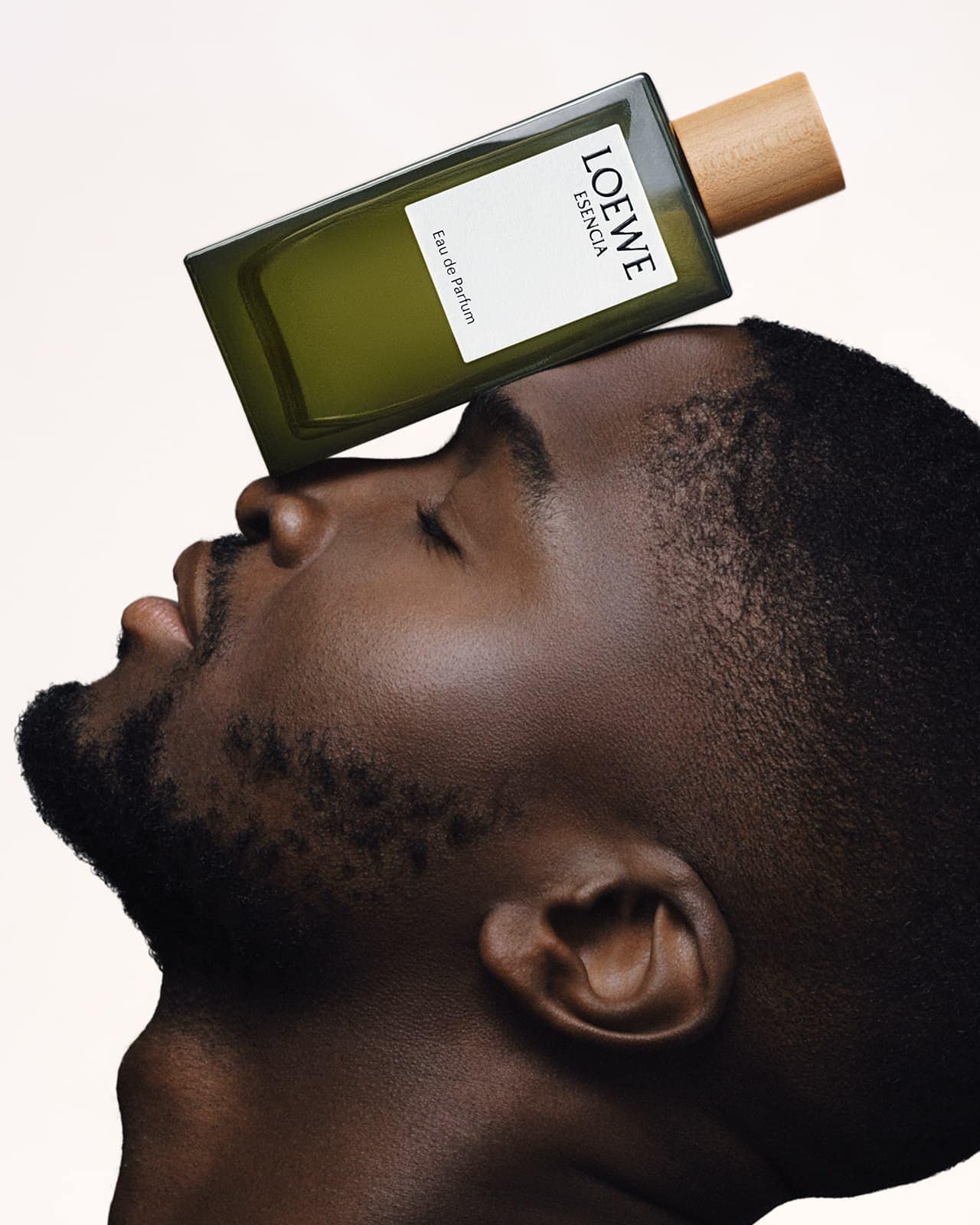 Masculine beauty shot of a Black male model with a Loewe perfume bottle balanced on his forehead against a neutral background.