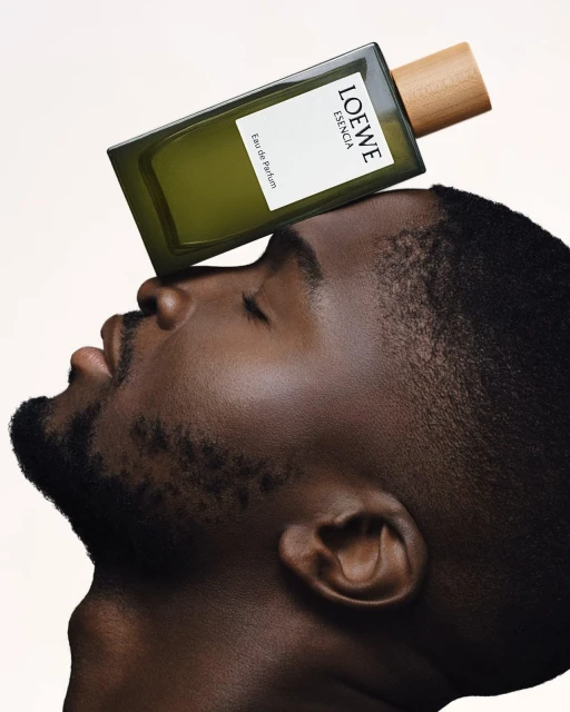 Masculine beauty shot of a Black male model with a Loewe perfume bottle balanced on his forehead against a neutral background.