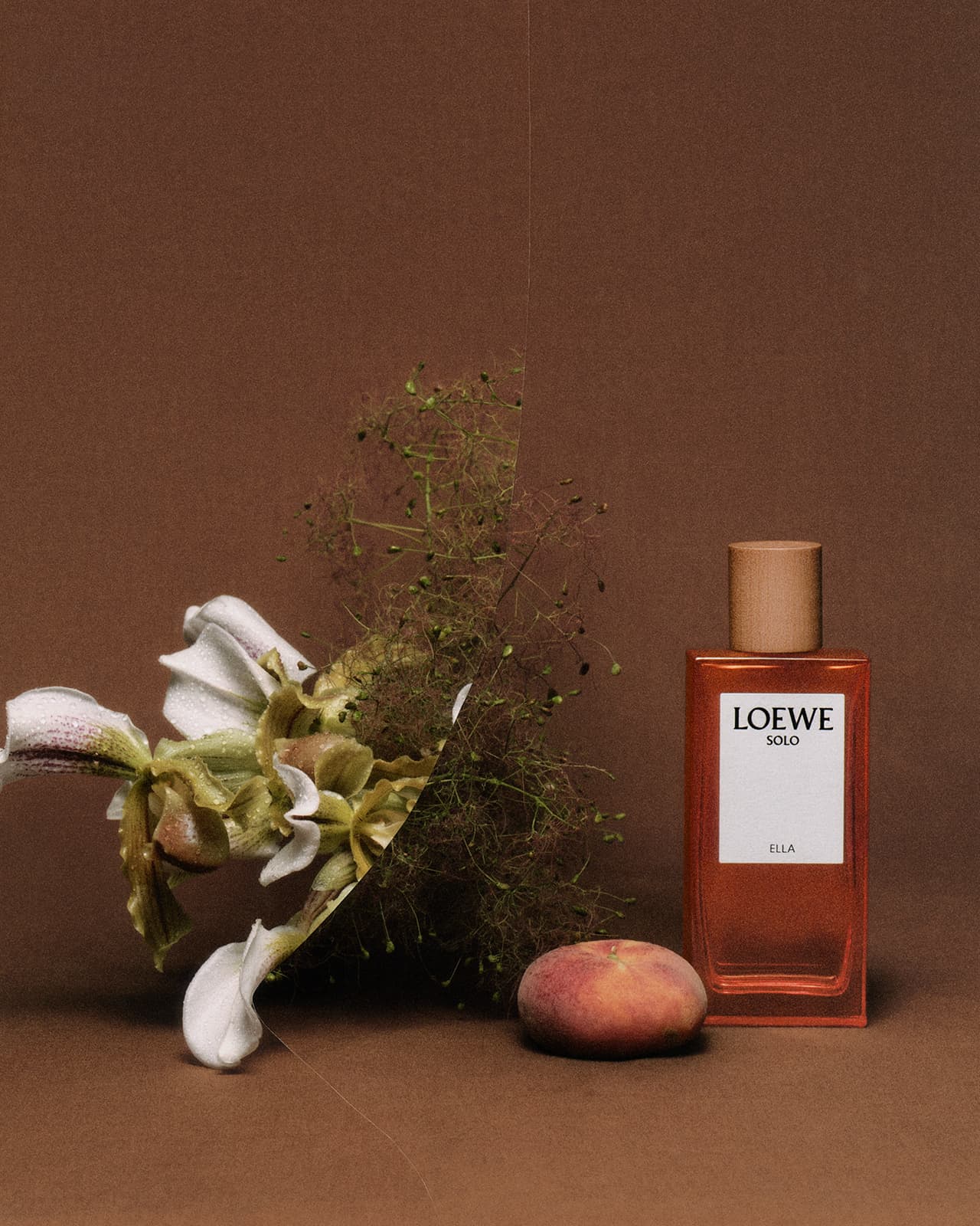 Still life shot of Loewe Solo Ella perfume bottle next to a peach and green and white flowers on a brown surface.