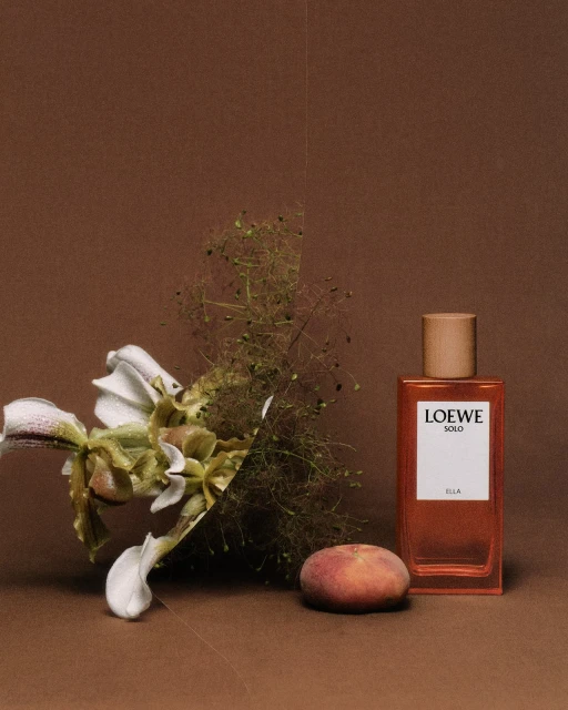 Still life shot of Loewe Solo Ella perfume bottle next to a peach and green and white flowers on a brown surface.