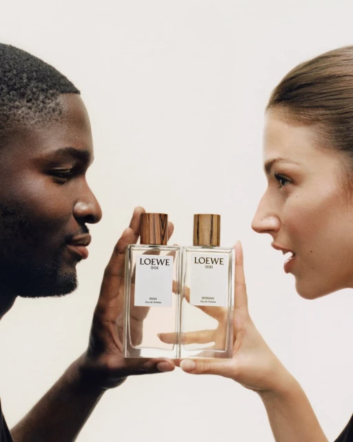 Loewe 001 perfume ad featuring an African-American male and Caucasian female holding two transparent bottles with gold caps.