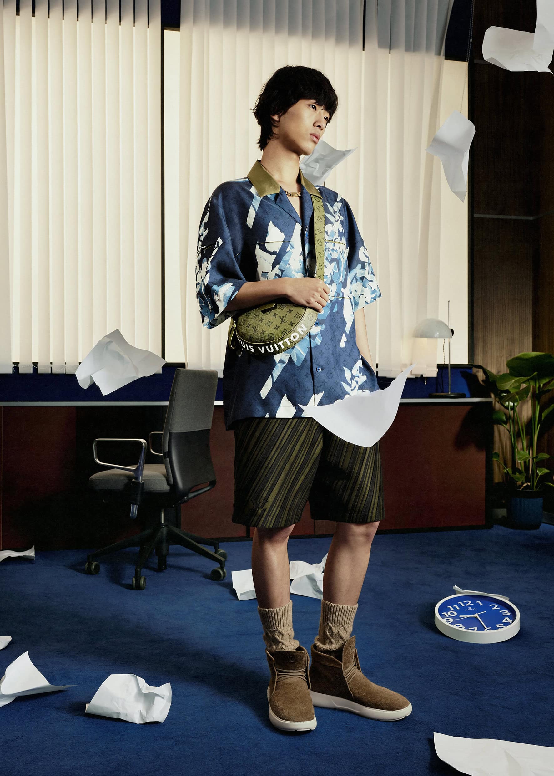 Stylish Asian man poses in an office with flying papers. He is wearing a patterned shirt, shorts, boots and a branded crossbody bag.