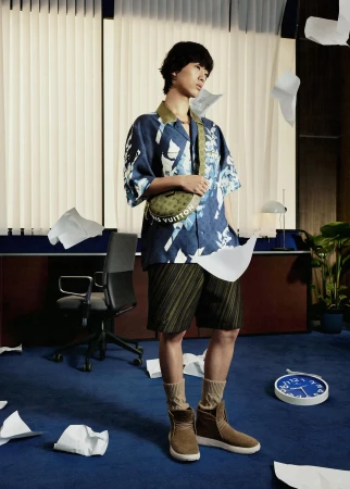 Stylish Asian man poses in an office with flying papers. He is wearing a patterned shirt, shorts, boots and a branded crossbody bag.