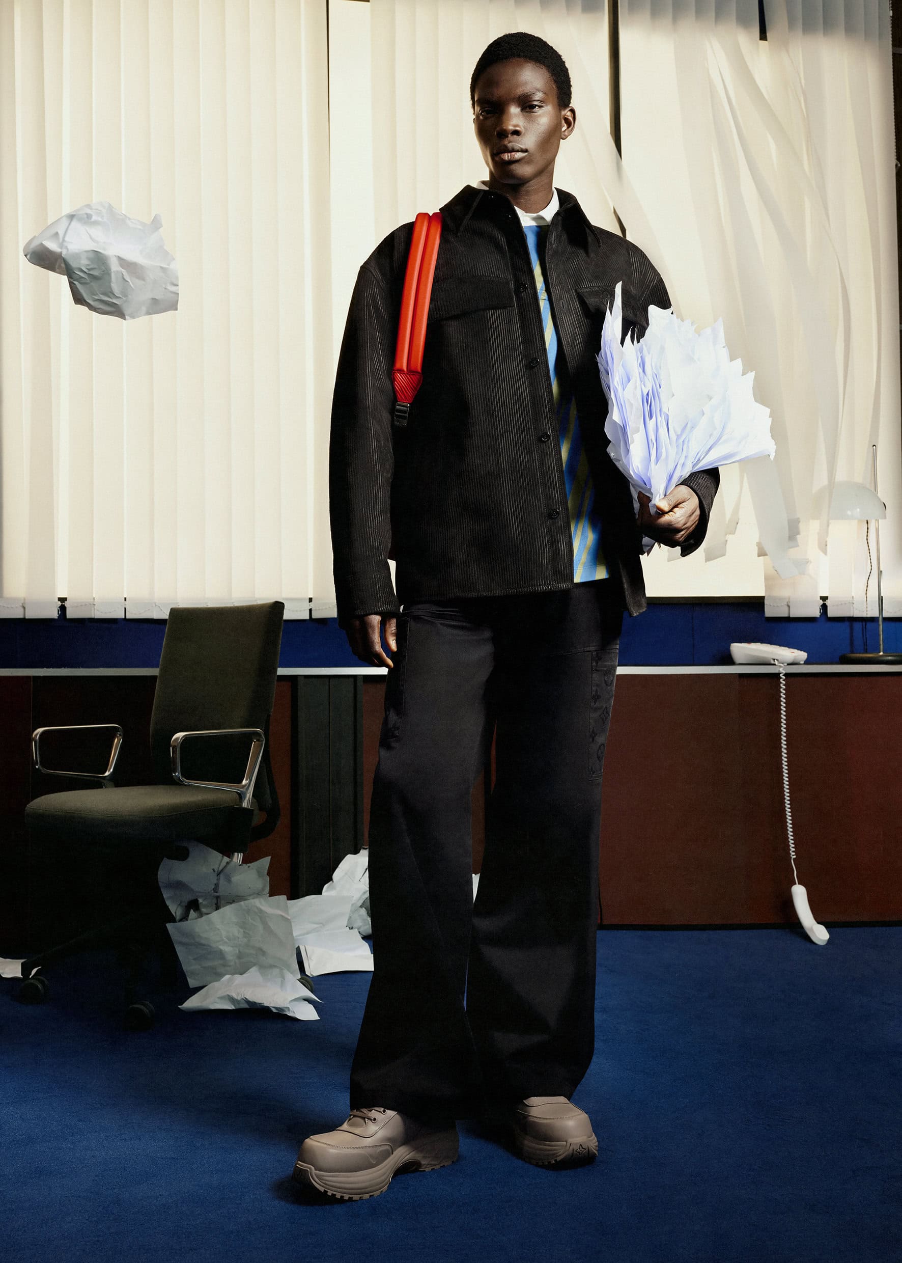A singular Black male model poses indoors in a black jacket, dark pants, and chunky tan sneakers with loose papers and a red backpack.