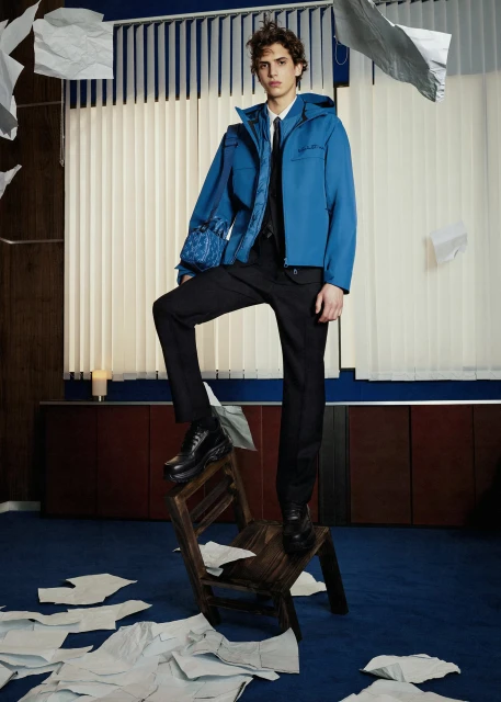 Male model in a blue jacket, black pants, and boots poses on a wooden step stool surrounded by scattered papers in an indoor setting.