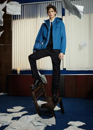Male model in a blue jacket, black pants, and boots poses on a wooden step stool surrounded by scattered papers in an indoor setting.