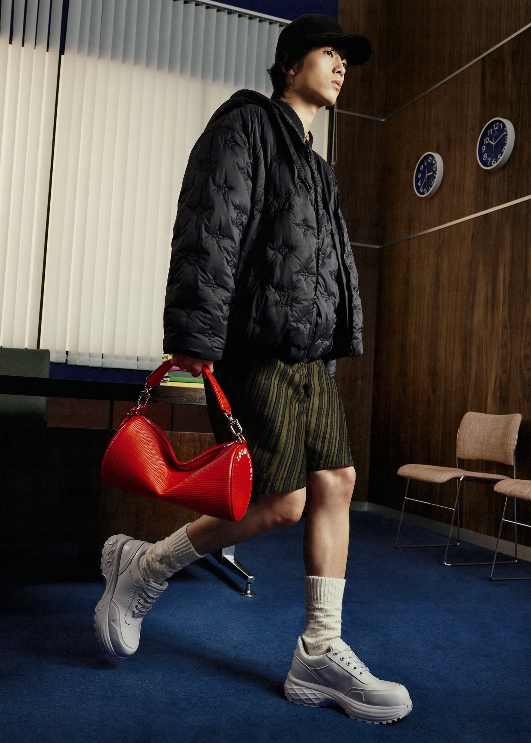 An Asian adult male models a black hooded jacket, green shorts, white sneakers, and a red handbag in an indoor studio setting.