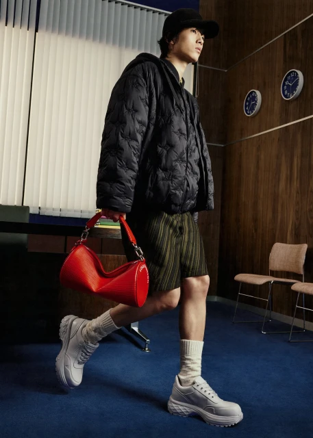 An Asian adult male models a black hooded jacket, green shorts, white sneakers, and a red handbag in an indoor studio setting.