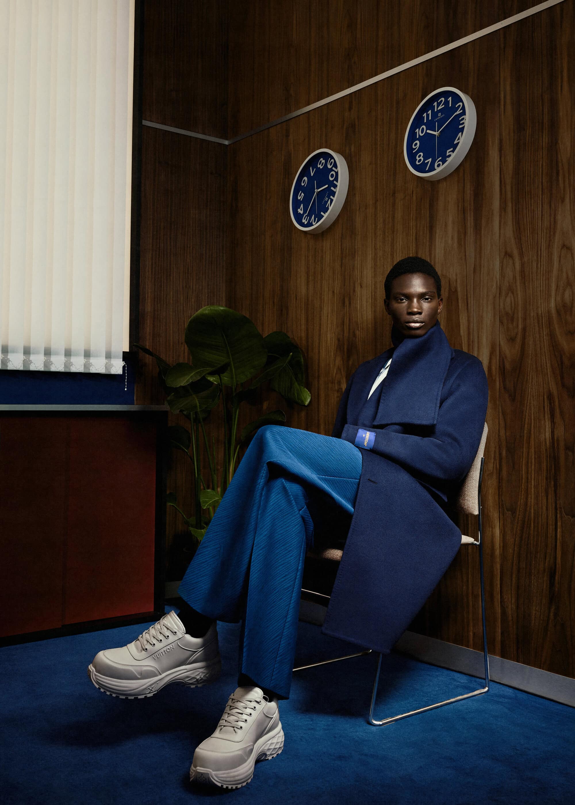 Dark-skinned male fashion model wearing blue coat and pants sitting in retro-style waiting room near blue analog clocks and green plant.