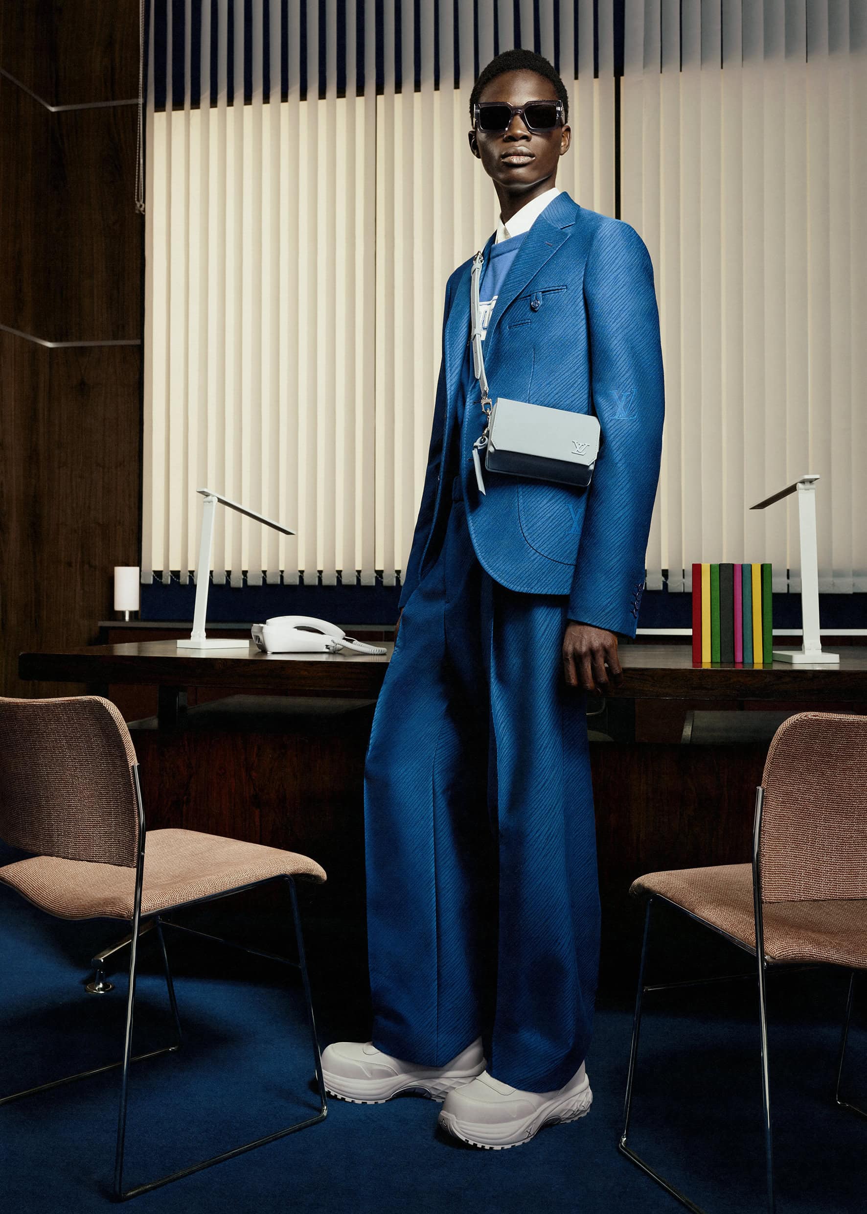 A stylish young Black man poses in a blue suit with a light-blue bag in an office setting with vertical blinds and desk lamps.
