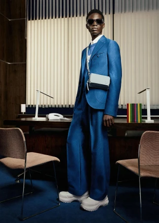 A stylish young Black man poses in a blue suit with a light-blue bag in an office setting with vertical blinds and desk lamps.