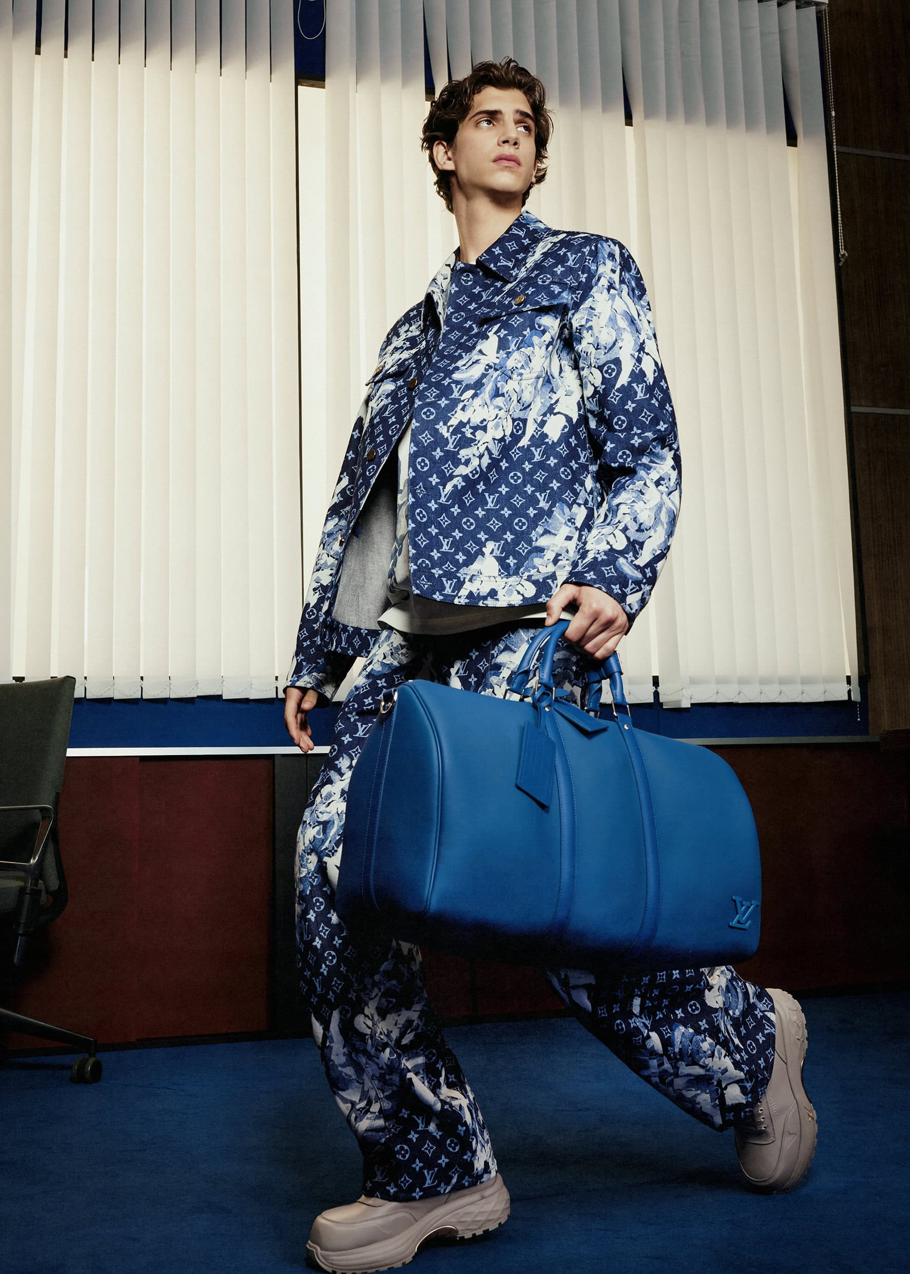 Adult male model in patterned jacket and pants carrying blue duffel bag in an office setting with vertical blinds and blue carpet.
