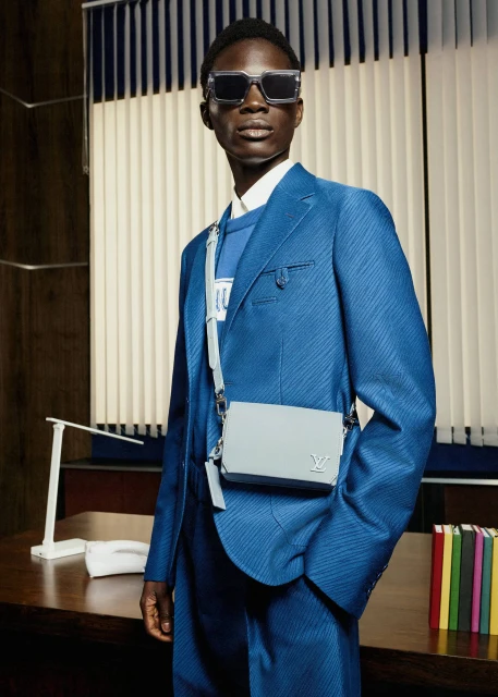 A Black male model wearing a blue suit and sunglasses stands in front of a striped window with a purse on his shoulder in a studio.