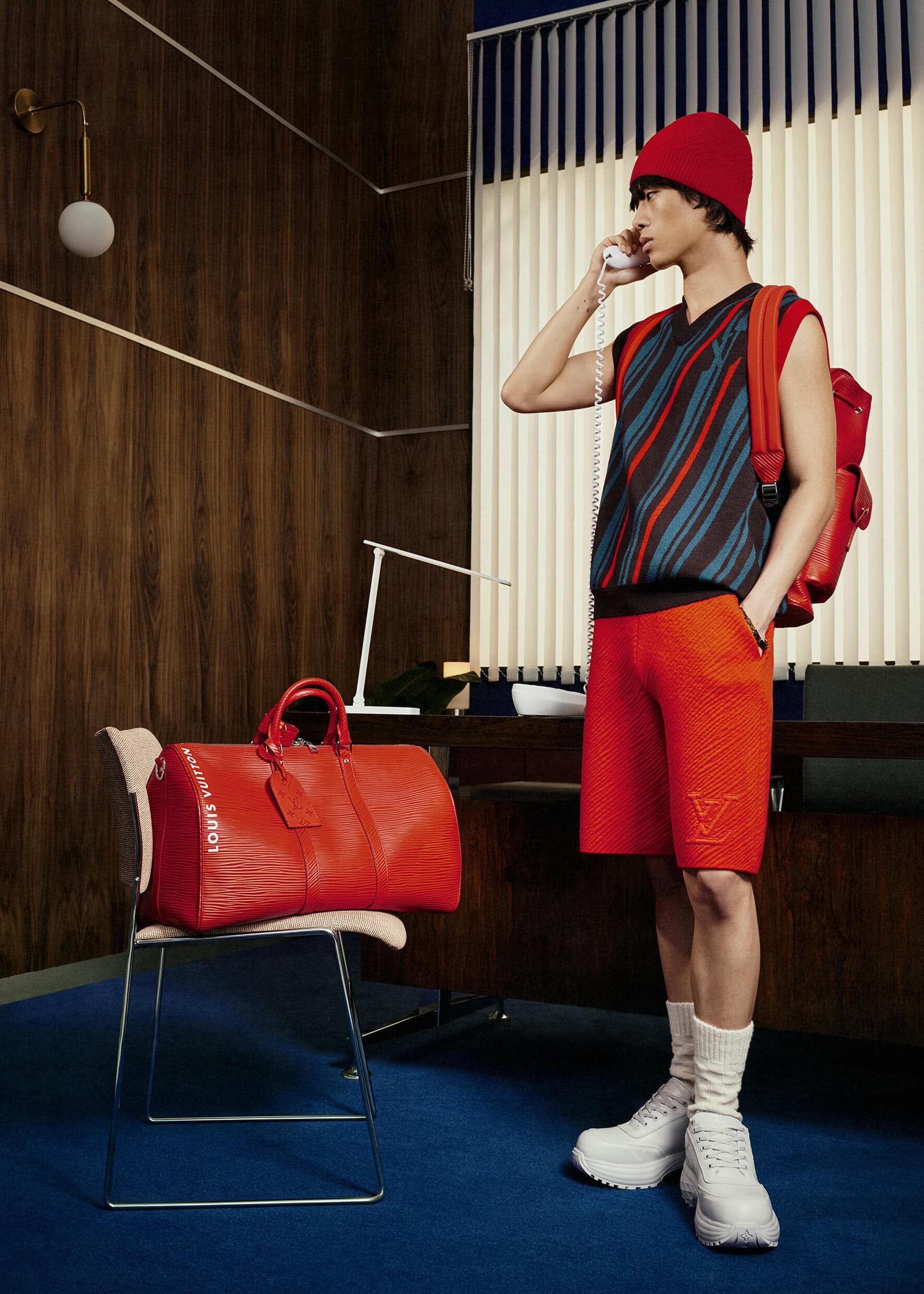 A man wearing a red beanie, striped shirt, and red shorts is talking on a phone in a room with wood paneling and blue carpet.