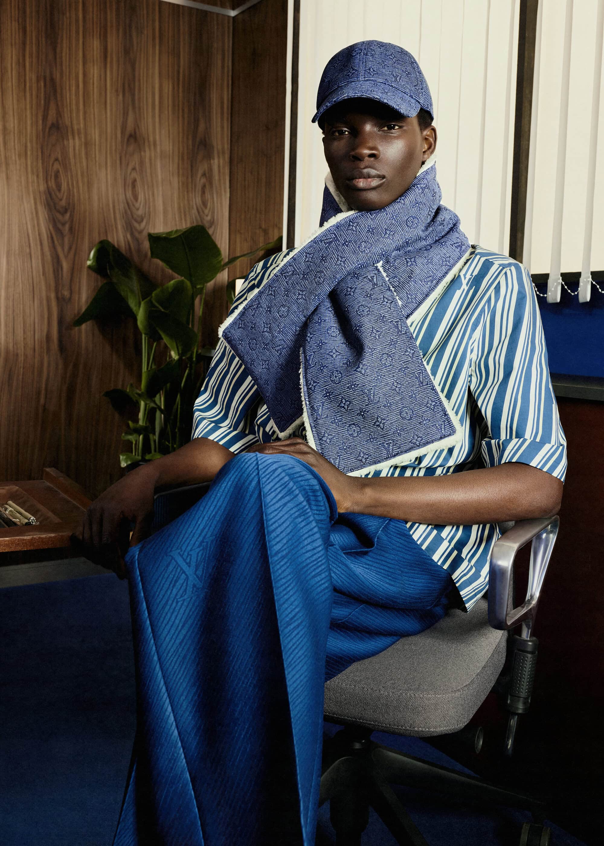 Dark-skinned man in blue striped shirt, cap, and scarf sits on an office chair in an indoor setting with a wood-paneled wall behind him.