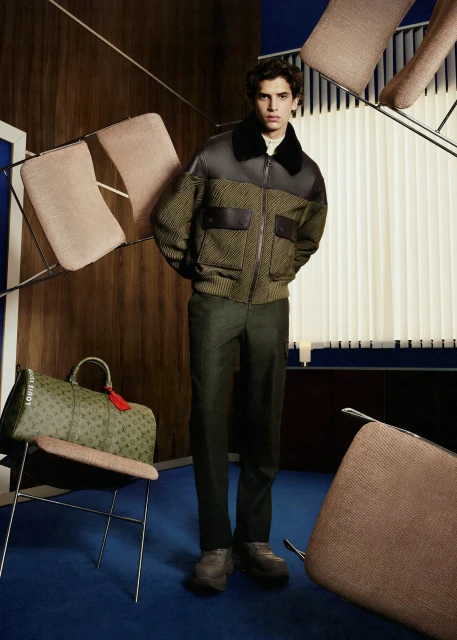 A stylish man in a bomber jacket poses indoors next to furniture, including chairs and a Louis Vuitton bag in an elegant fashion studio.