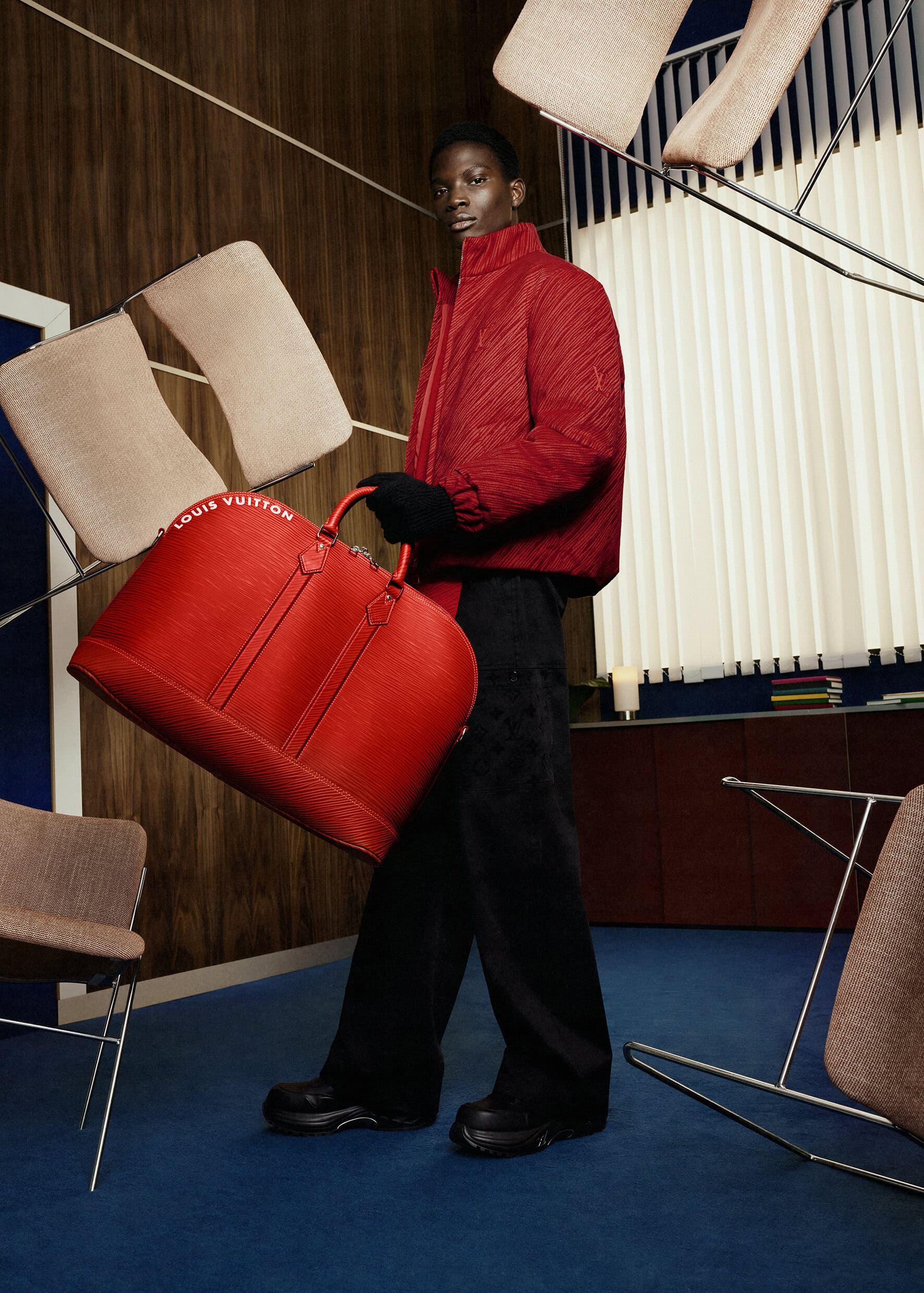 An adult male model poses with a red Louis Vuitton bag in a studio setting with blue floors, wooden paneling, and suspended chair parts.