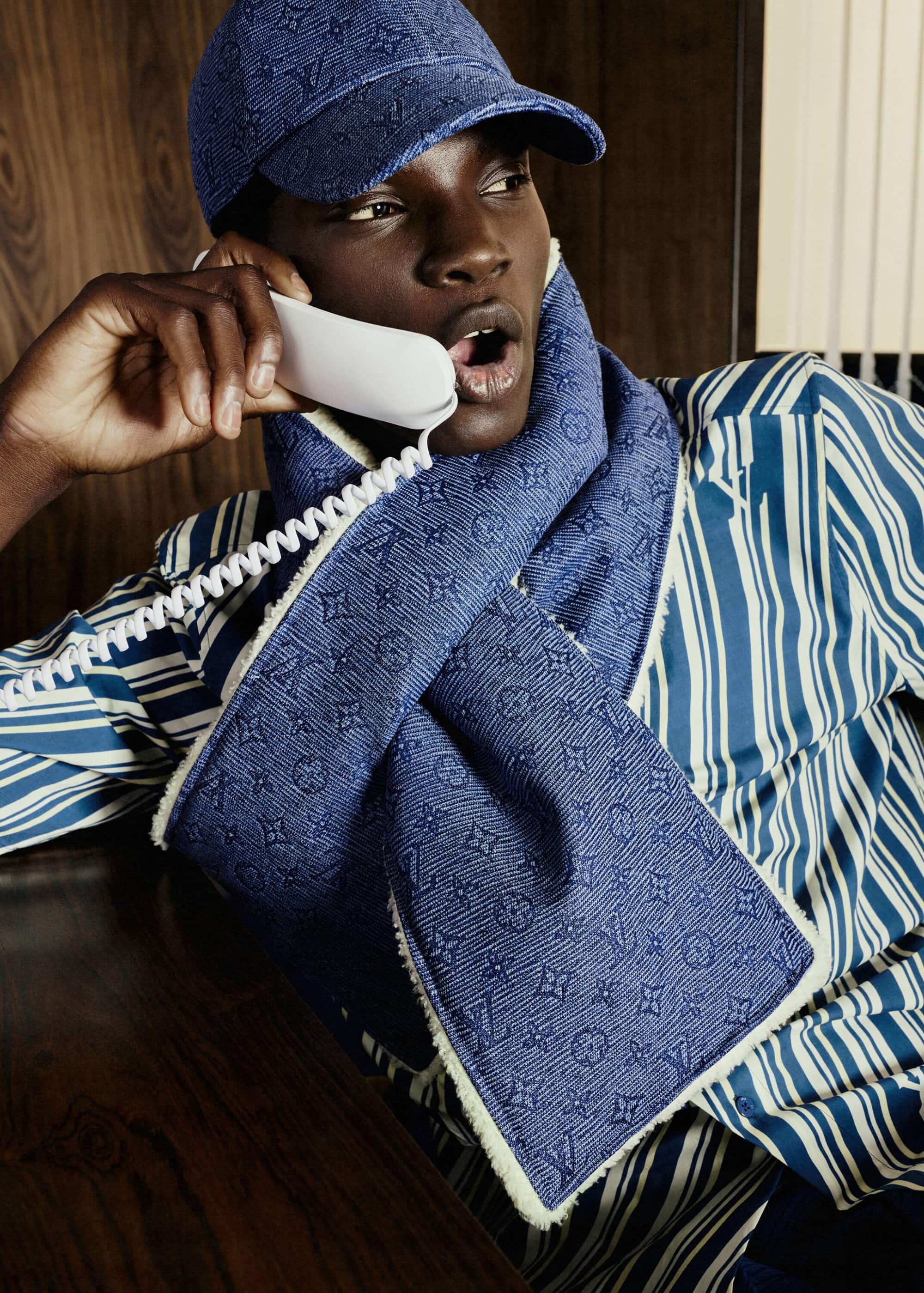 Person in baseball cap and monogrammed scarf talking on a retro telephone with a striped shirt in a wood paneled room.