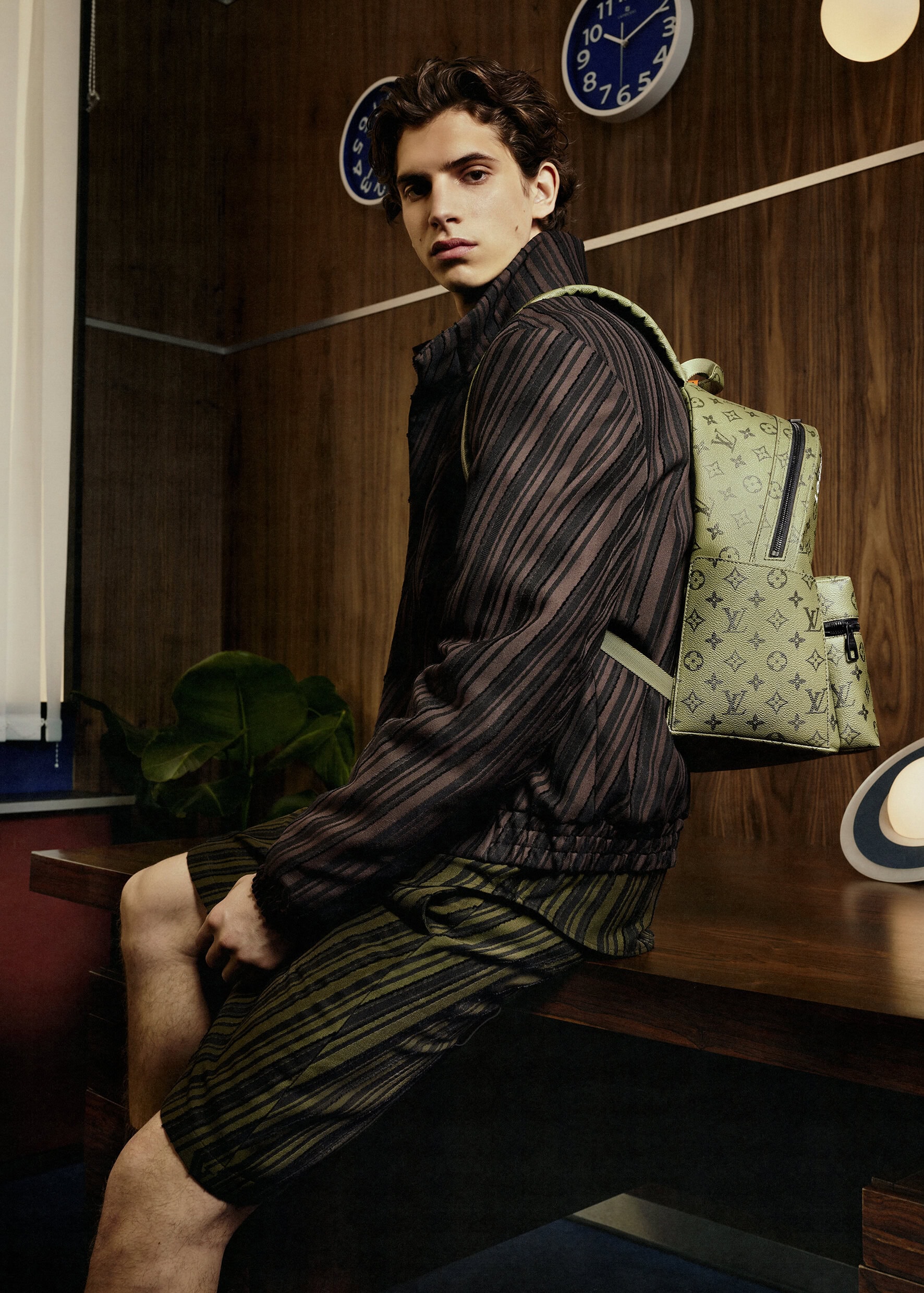 Young adult man in patterned shorts and jacket with a backpack sits on a desk in front of a wood paneled wall with clocks.
