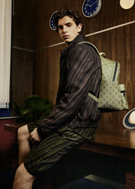 Young adult man in patterned shorts and jacket with a backpack sits on a desk in front of a wood paneled wall with clocks.