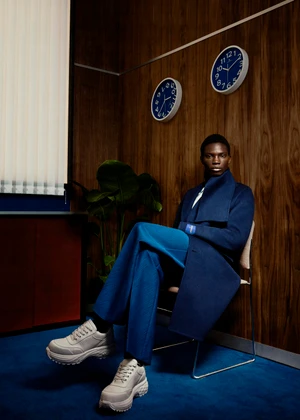 Black male model seated in a navy coat, blue trousers, and chunky sneakers against a wood-paneled office wall with two clocks.