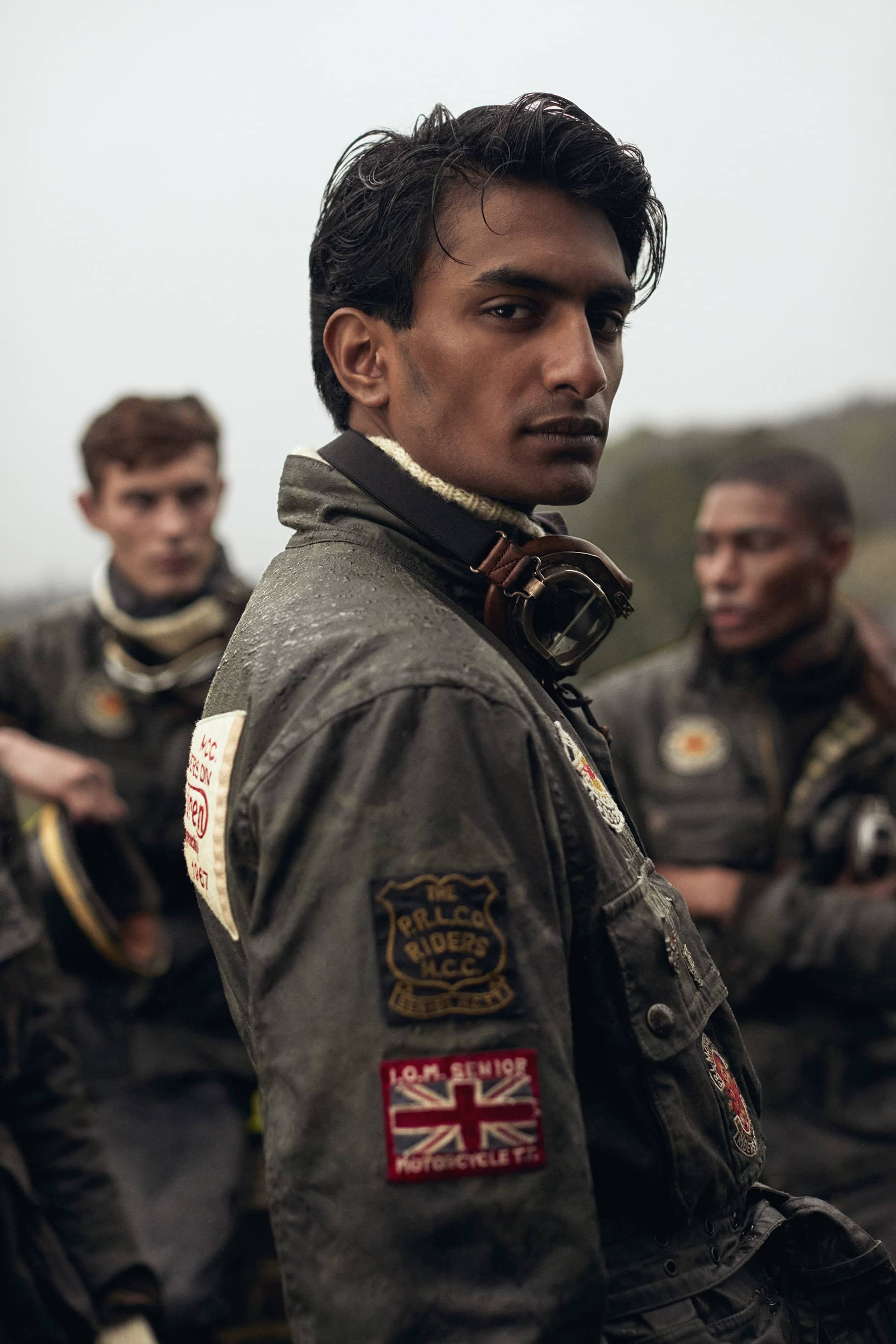 A group of three men in vintage racing attire are standing in an outdoor setting with focus on the man in the foreground looking back.