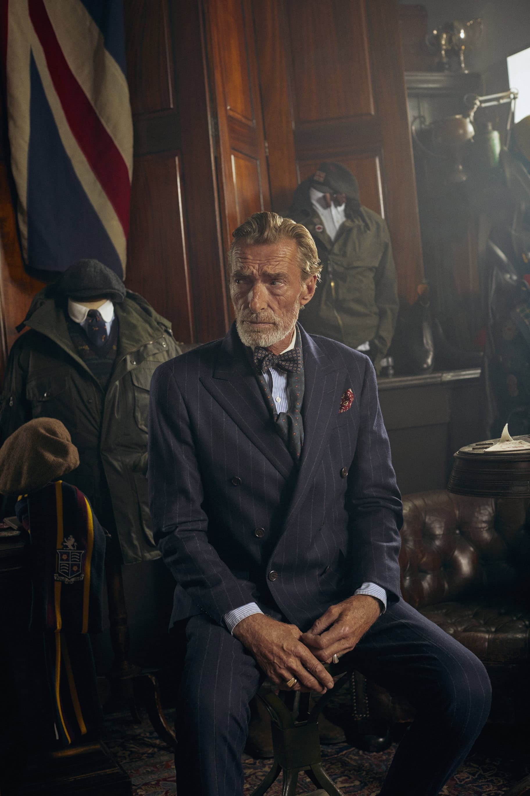 Mature man in a pinstripe suit and bow tie sits in a dimly lit indoor setting near a Union Jack flag and clothing mannequins.