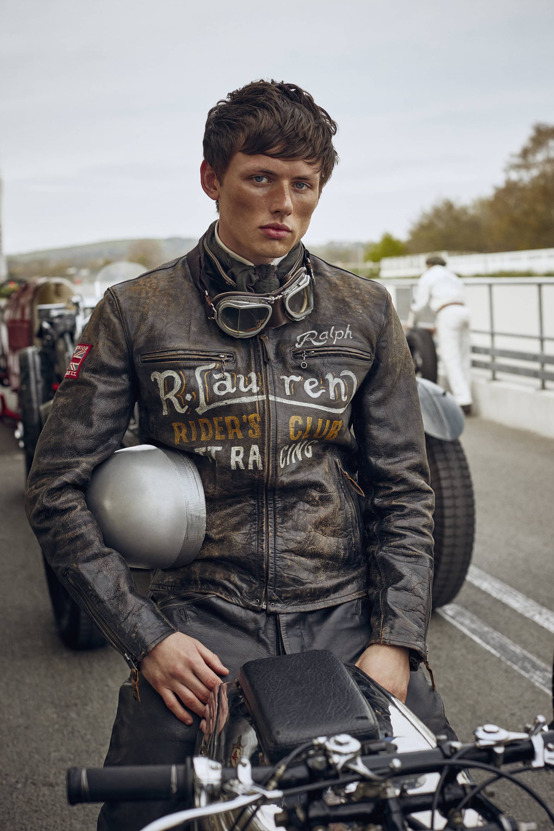 Man in a distressed leather jacket sits on a vintage motorcycle with a silver helmet and goggles, outdoors at a racetrack.