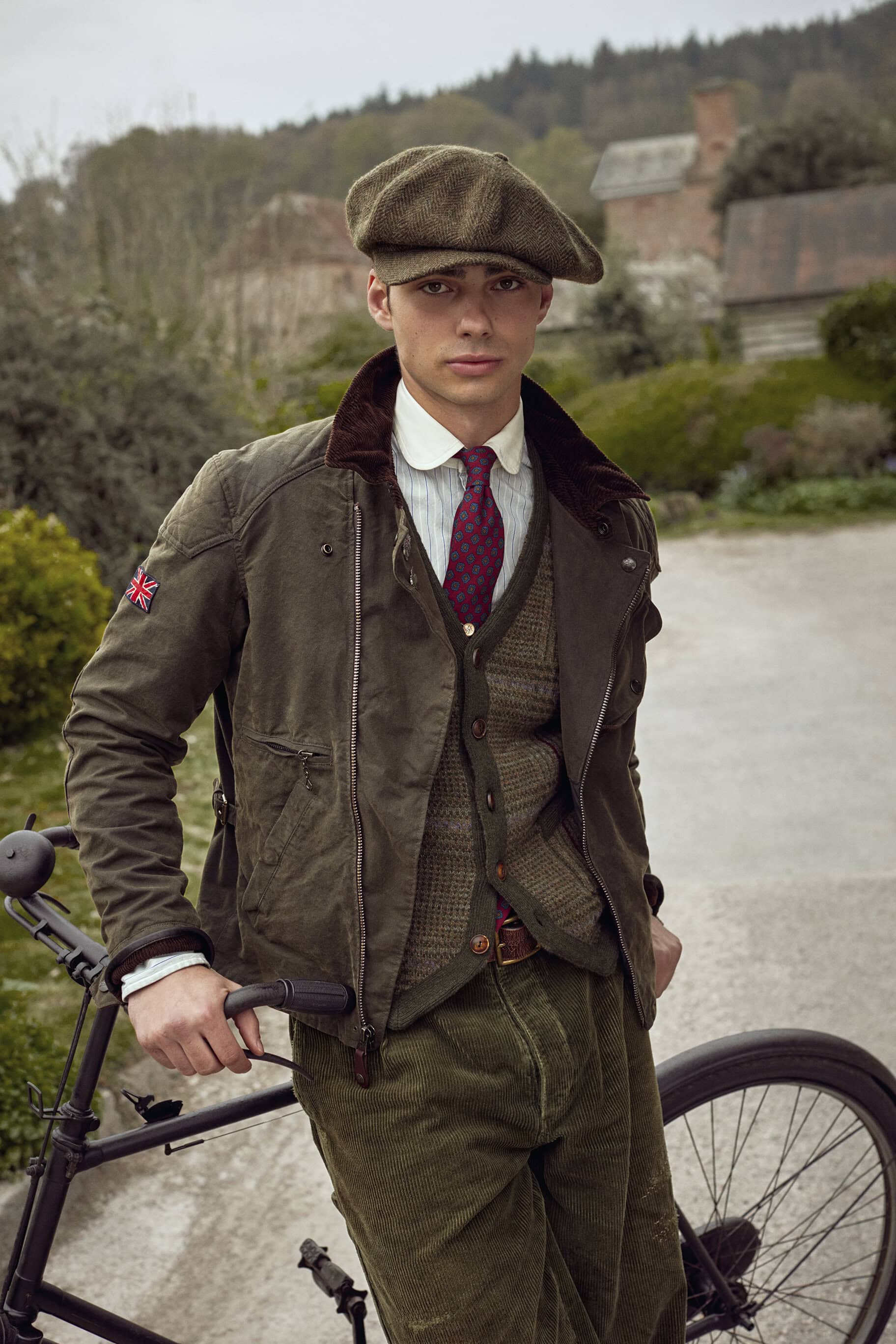 Male model in classic British attire and cap poses with vintage bicycle on a rural road with a cottage in the background.