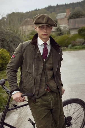 Male model in classic British attire and cap poses with vintage bicycle on a rural road with a cottage in the background.