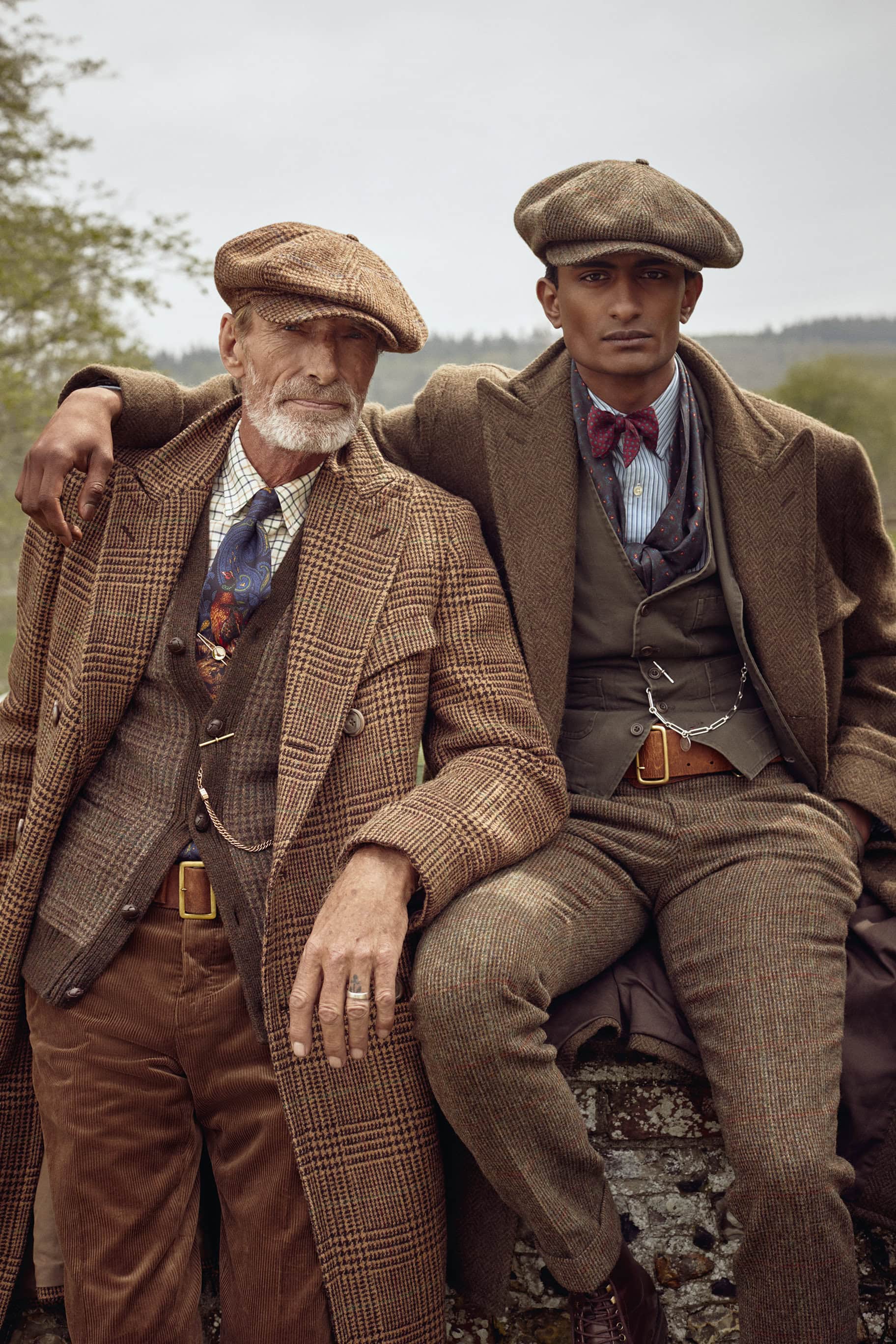 Two men in tweed suits and caps pose outdoors; one seated on a stone wall with arm around the other, creating a vintage fashion portrait.