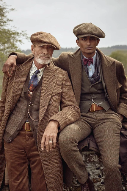 Two men in tweed suits and caps pose outdoors; one seated on a stone wall with arm around the other, creating a vintage fashion portrait.