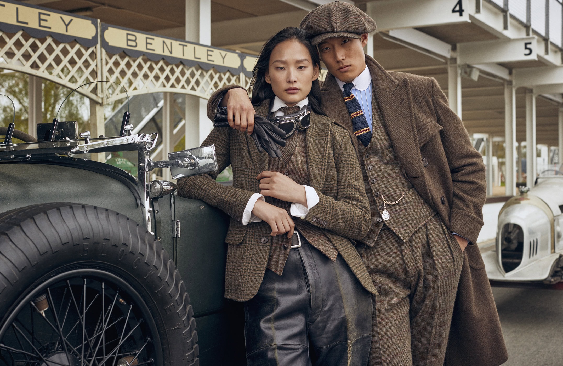 A fashionable couple poses with a vintage Bentley race car on a racing track, wearing tweed suits and caps in a vintage-inspired fashion shoot.
