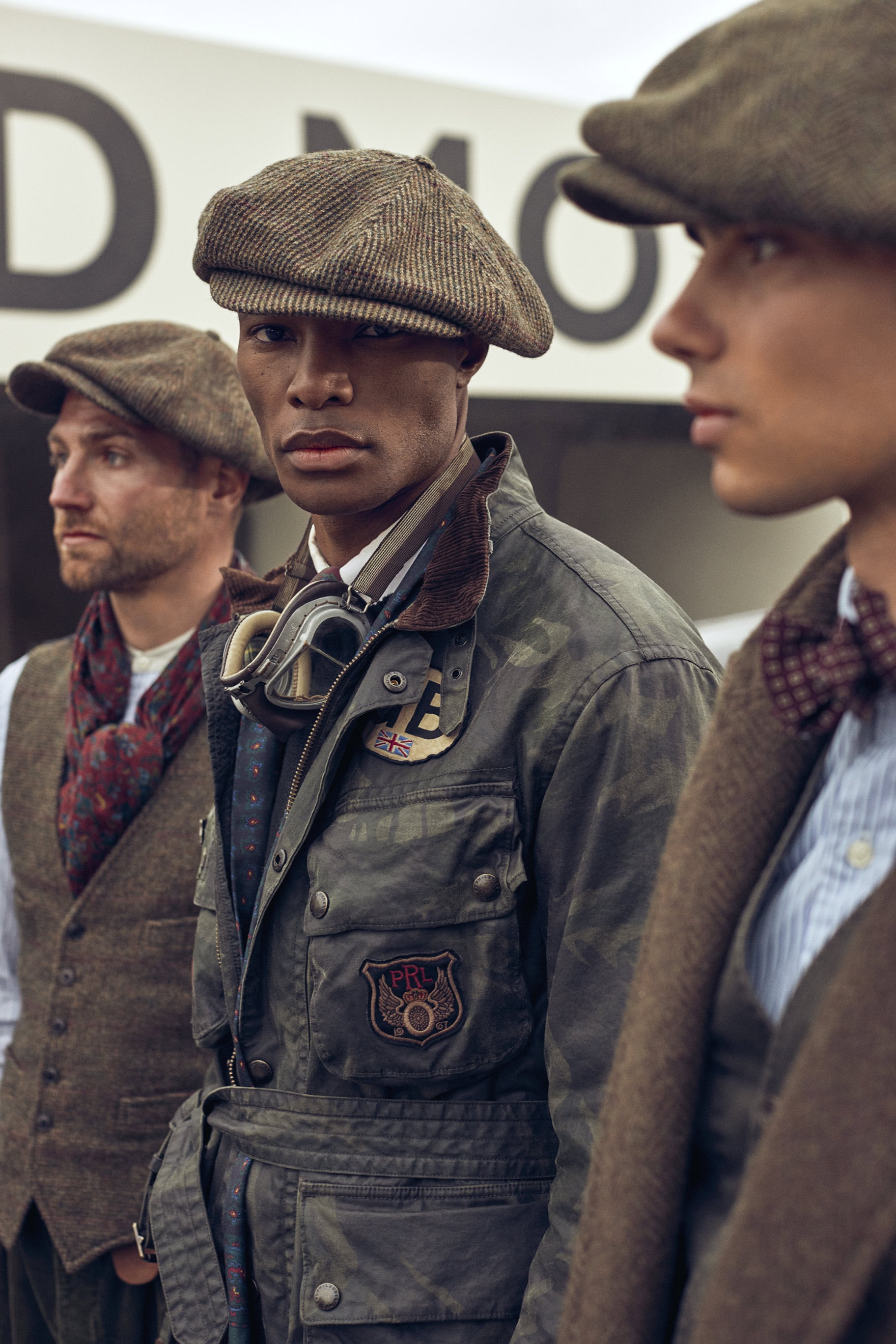Three stylish adult males stand in a row wearing vintage-inspired clothing, caps, and goggles, creating a retro fashion statement.