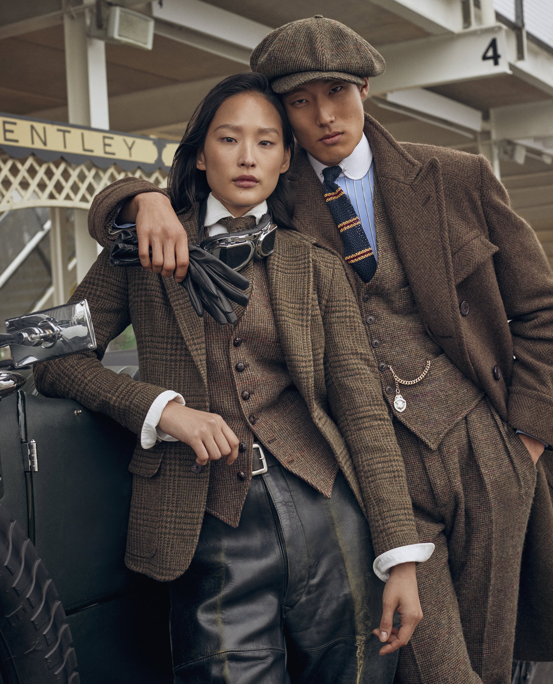 A stylish couple poses outdoors in brown tweed suits with vintage leather accents, evoking a classic early 20th-century automobile aesthetic.