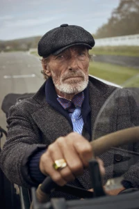 Elderly bearded man in a charcoal tweed flat cap and herringbone jacket gripping the steering wheel of a vintage open-top car.